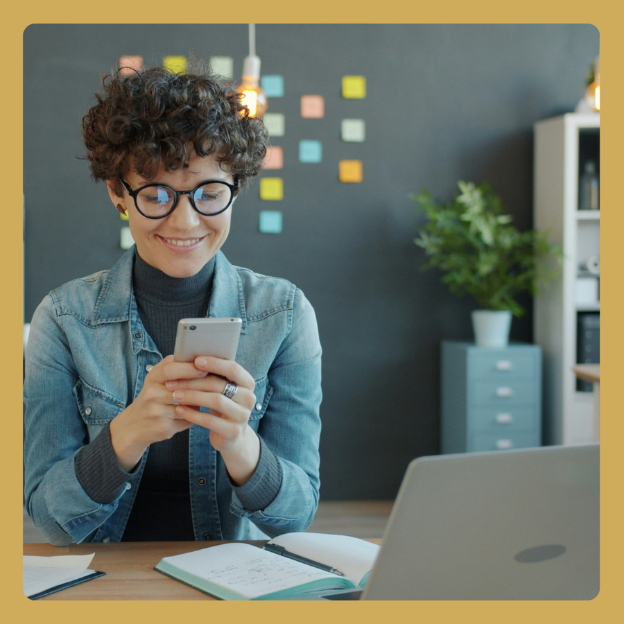 Person with curly hair and glasses using smartphone at desk with laptop, notebook, sticky notes