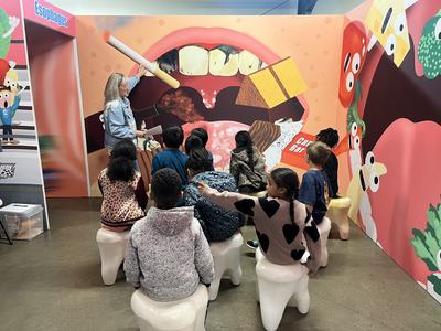 Teacher points to mural of open mouth as children sit on tooth-shaped stools; "Esophagus" sign visible.