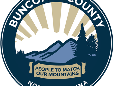 County government logo for Buncombe County. A graphic of blue mountains, dark blue pine trees, and brown sun rays with white clouds over the mountains.