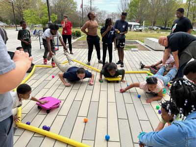 Kids on scooter boards reaching for colorful balls as adults hold their legs on an outdoor plaza