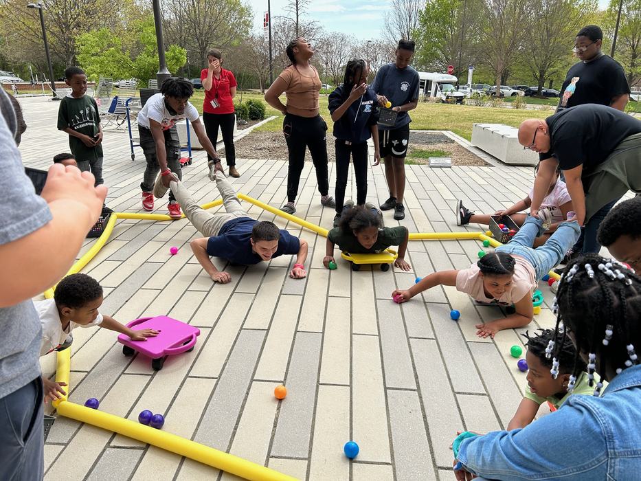 Kids on scooter boards reaching for colorful balls as adults hold their legs on an outdoor plaza