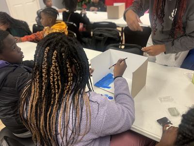 Child painting blue inside a white cardboard diorama box at a classroom table