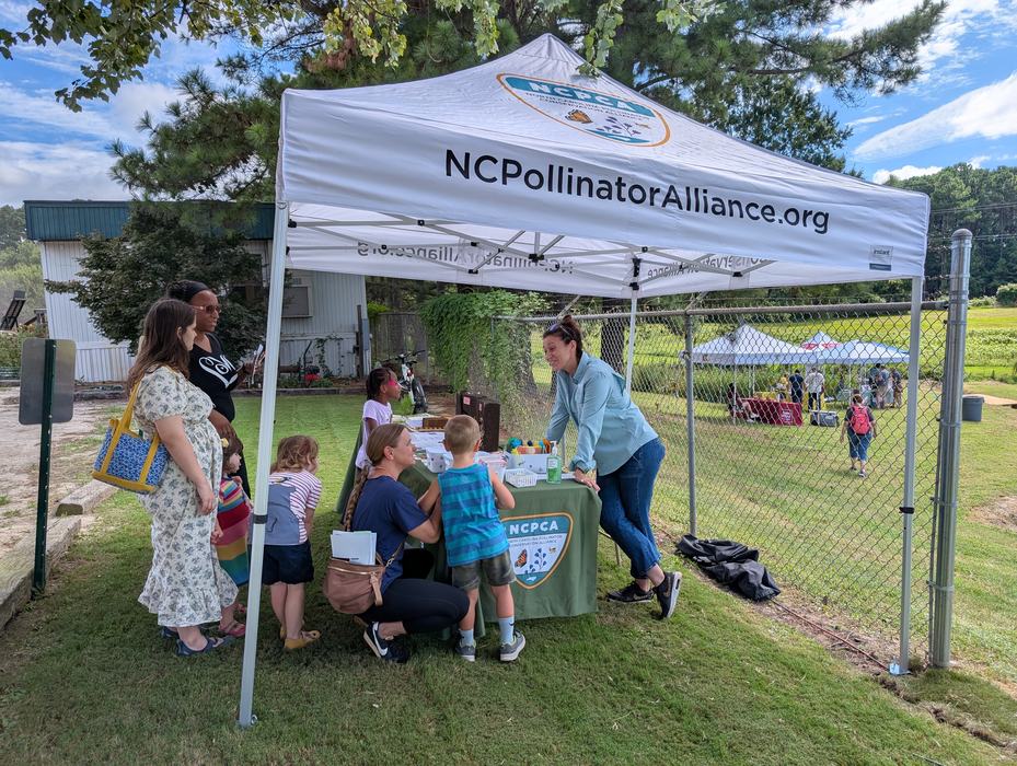A member of the North Carolina Pollinator Conservation Alliance talks to a family about pollinators and the activities at the NC Pollinator Field Day at the State Farmers Market in Raleigh