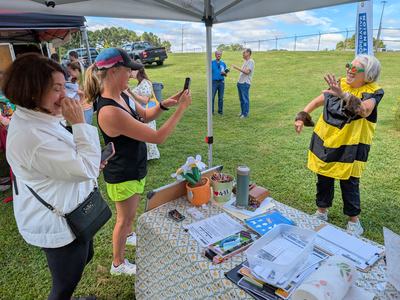 Person in yellow-black bee costume posing under tent while woman photographs them at an outdoor fair