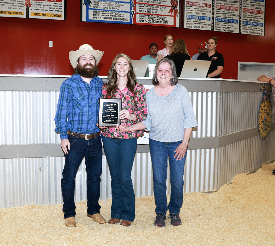Three people standing in an auction arena; center woman holds a plaque