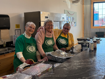 Three older adults in green aprons behind a counter serving food trays in a community kitchen