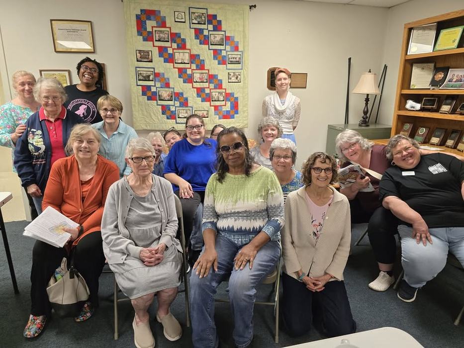 A group photo of ECA members with a memory quilt hanging as a back drop.