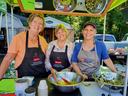 Three women in aprons serving salad at an outdoor food volunteer booth