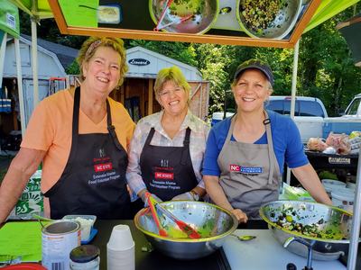 Three women in aprons serving salad at an outdoor food volunteer booth