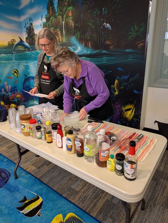 Two women wearing NC STATE aprons setting up condiments and utensils on a table