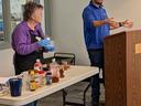 Woman wearing gloves arranges condiments on table while man speaks at podium