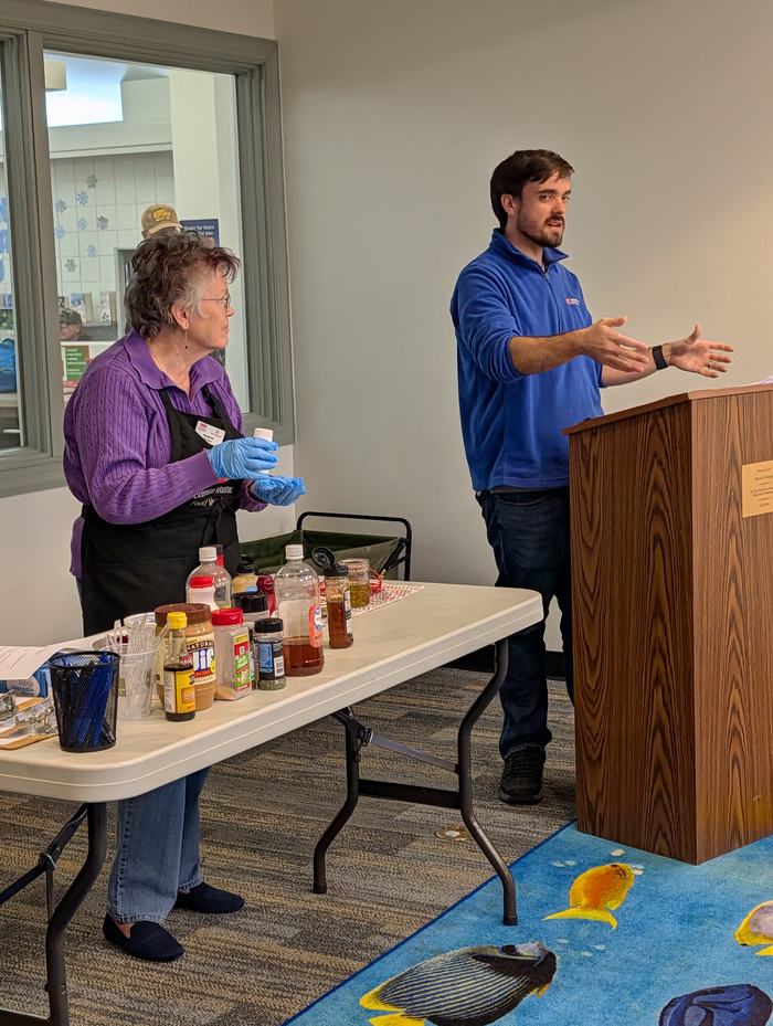 Woman wearing gloves arranges condiments on table while man speaks at podium