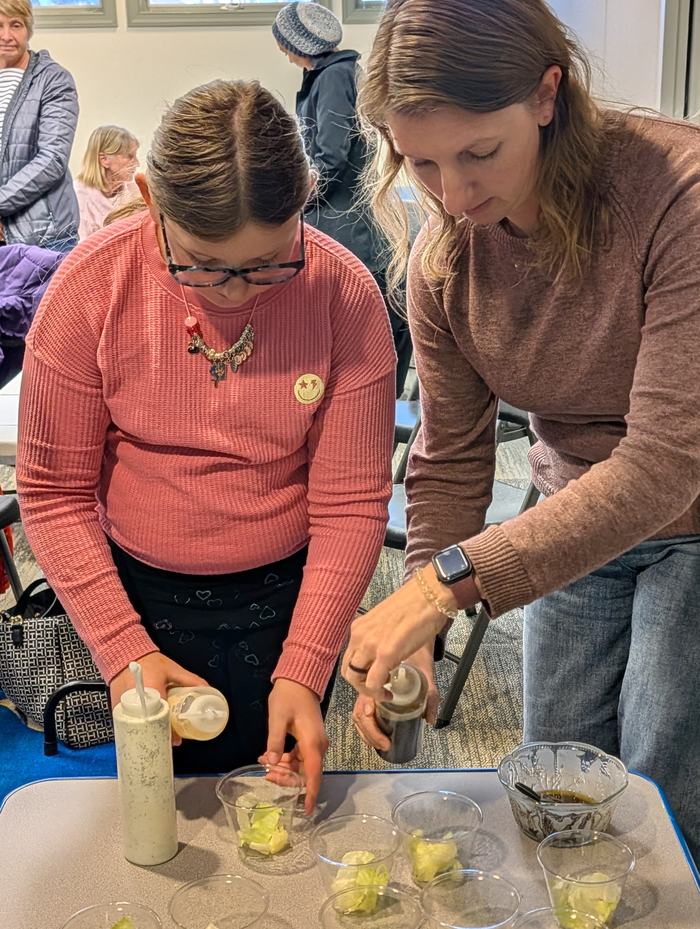 Young girl and adult woman pouring dressing into plastic cups with lettuce