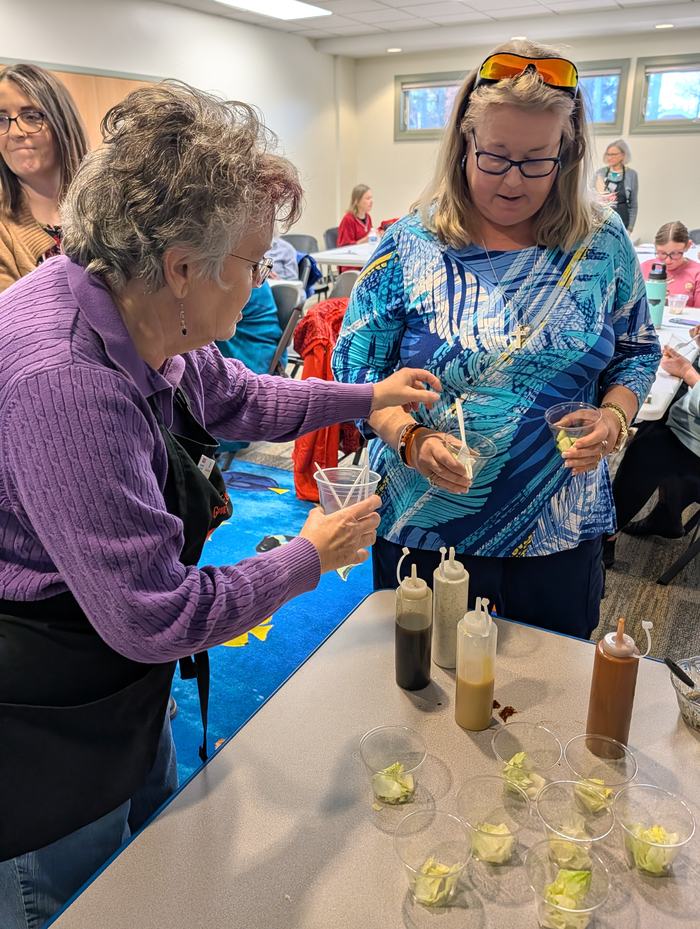 Two women preparing salad samples at a table with squeeze bottles and plastic cups