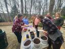 A group of people standing around a table full of pots of media, cutting boards, knives, and roots of various native woodland botanicals. This was a workshop to teach propagation.