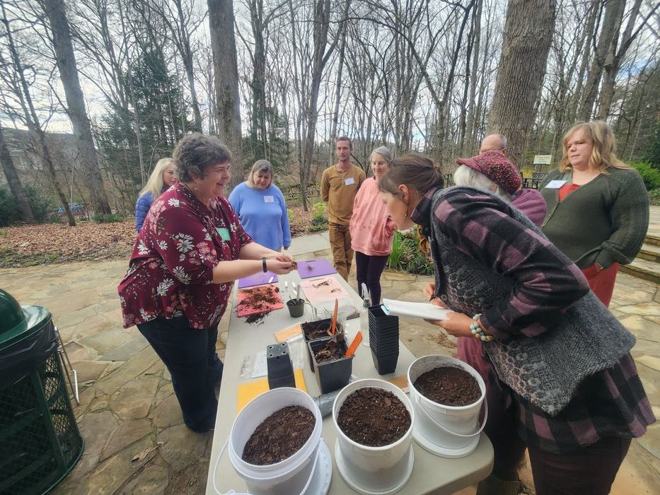 A group of people standing around a table full of pots of media, cutting boards, knives, and roots of various native woodland botanicals. This was a workshop to teach propagation.