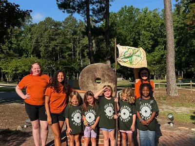 Camp staff and children posing in front of a large stone wheel, one child holding a flag
