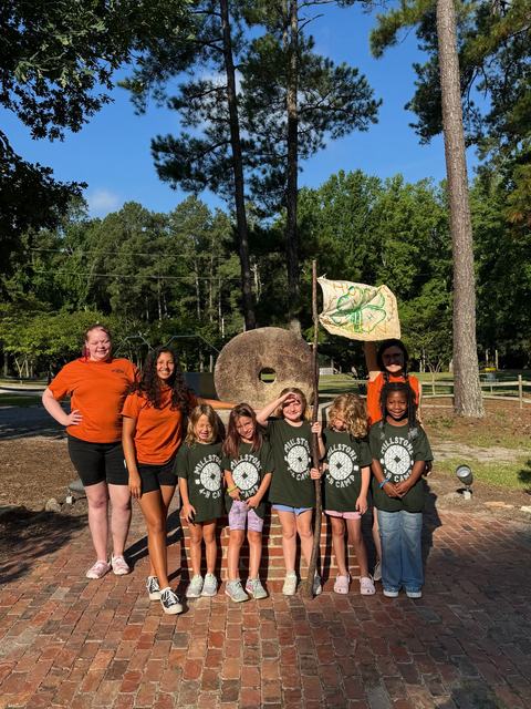 Camp staff and children posing in front of a large stone wheel, one child holding a flag