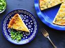Top-down view of a golden-brown broccoli and cheese quiche in a blue ruffled pie dish, with one slice served on a matching decorative blue plate next to a silver fork and fresh broccoli.