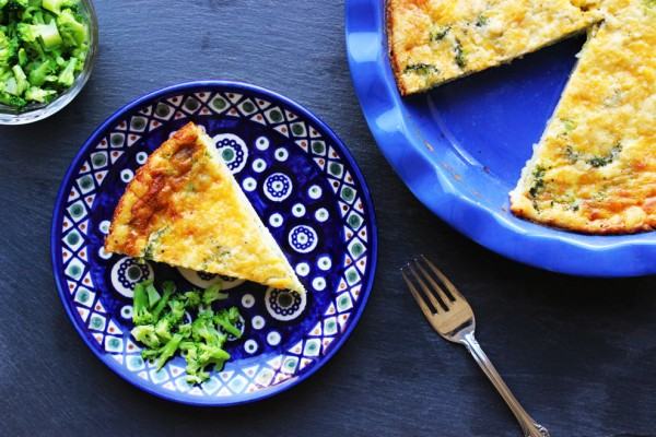 Top-down view of a golden-brown broccoli and cheese quiche in a blue ruffled pie dish, with one slice served on a matching decorative blue plate next to a silver fork and fresh broccoli.