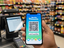 Hand holding a smartphone displaying a rewards program QR code at a supermarket checkout counter.