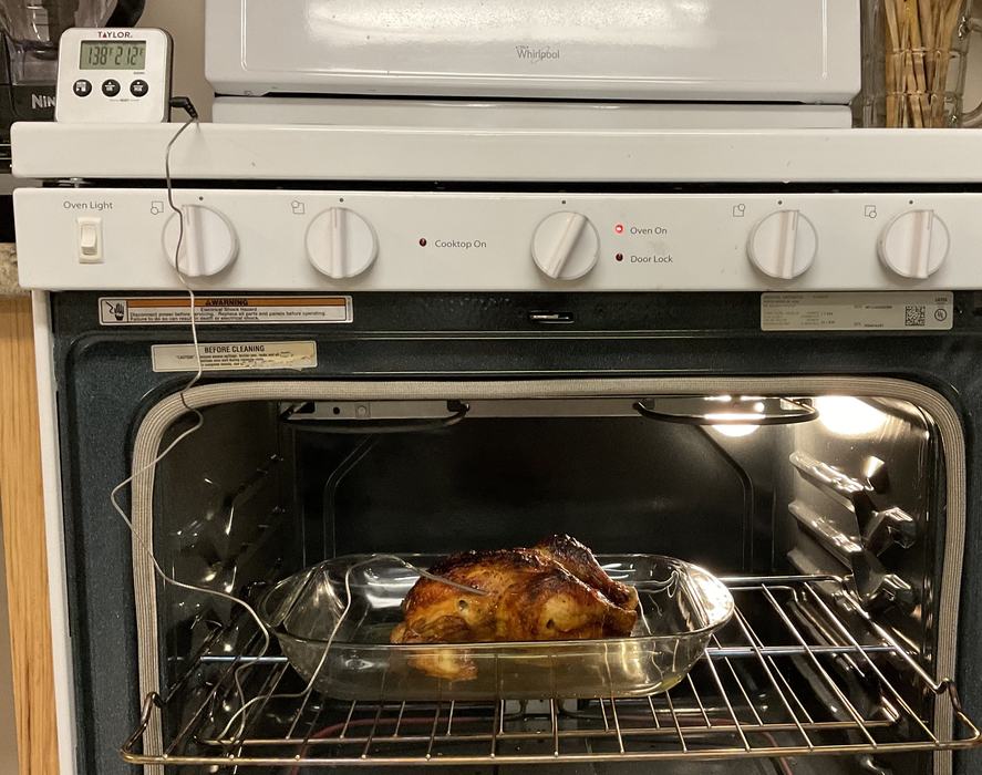 Cooked chicken in a glass baking dish in an oven with an open door with an oven cord thermometer inserted into it and the base of the thermometer sitting on top of the white stove.