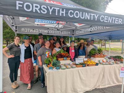 Group of vendors under "Forsyth County Center" tent at farmers market with baskets of produce.