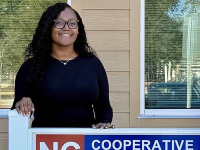 A WOMAN STANDING IN FRONT OF A SIGN THAT READS NC COOPERATIVE EXTENSION BERTIE COUNTY WITH NC A&T AND NC STATE LOGOS ON SIGN.