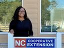 A WOMAN STANDING IN FRONT OF A SIGN THAT READS NC COOPERATIVE EXTENSION BERTIE COUNTY WITH NC A&T AND NC STATE LOGOS ON SIGN.