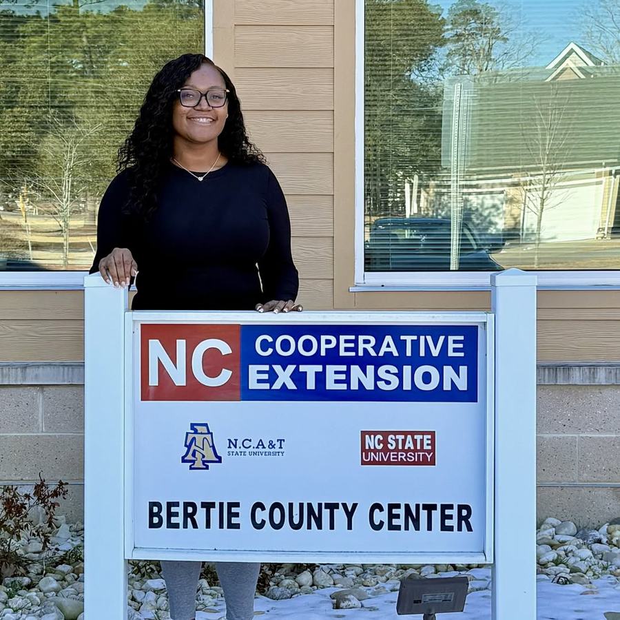 A WOMAN STANDING IN FRONT OF A SIGN THAT READS NC COOPERATIVE EXTENSION BERTIE COUNTY WITH NC A&T AND NC STATE LOGOS ON SIGN.