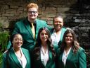 The five 2026-2027 State 4-H Council officers wearing green jackets pose together in front of a stone wall, greenery and a waterfall.
