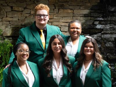 The five 2026-2027 State 4-H Council officers wearing green jackets pose together in front of a stone wall, greenery and a waterfall.