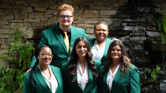 The five 2026-2027 State 4-H Council officers wearing green jackets pose together in front of a stone wall, greenery and a waterfall.
