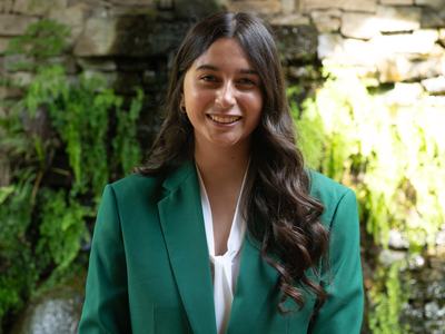 Natalia Estrada, 2026-2027 State 4-H Council President, wearing a green jacket and white blouse, standing in front of a stone wall with greenery and a waterfall.