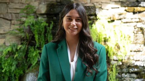 Natalia Estrada, 2026-2027 State 4-H Council President, wearing a green jacket and white blouse, standing in front of a stone wall with greenery and a waterfall.