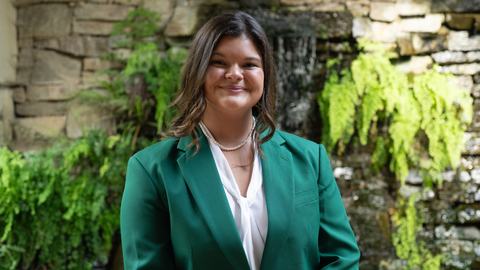 Leah Frick, 2026-2027 State 4-H Council Vice President, wearing a green jacket and white blouse, standing in front of a stone wall with greenery and a waterfall.