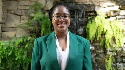 Promise McCoy, 2026-2027 State 4-H Council Secretary-Treasurer, wearing a green jacket and white blouse, standing in front of a stone wall with greenery and a waterfall.