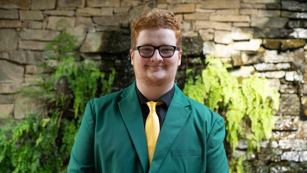 Kendal Thornburg, 2026-2027 State 4-H Council Member Engagement, wearing a green jacket with a black shirt and gold tie, standing in front of a stone wall with greenery and a waterfall.