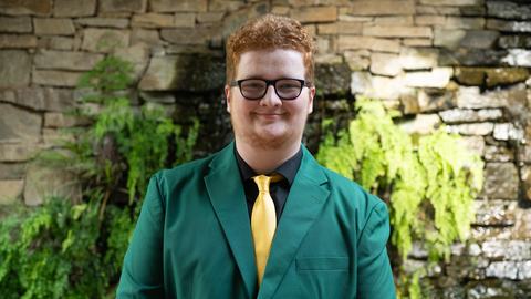 Kendal Thornburg, 2026-2027 State 4-H Council Member Engagement, wearing a green jacket with a black shirt and gold tie, standing in front of a stone wall with greenery and a waterfall.