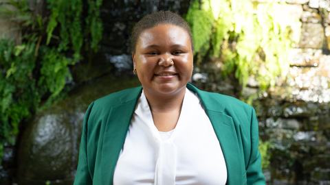Galyce Ham, 2026-2027 State 4-H Council Reporter, wearing a green jacket and white blouse, standing in front of a stone wall with greenery and a waterfall.