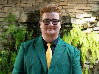 Kendal Thornburg, 2026-2027 State 4-H Council Member Engagement, wearing a green jacket with a black shirt and gold tie, standing in front of a stone wall with greenery and a waterfall.