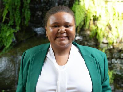 Galyce Ham, 2026-2027 State 4-H Council Reporter, wearing a green jacket and white blouse, standing in front of a stone wall with greenery and a waterfall.