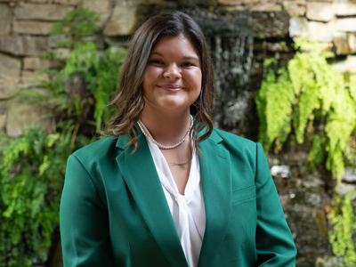 Leah Frick, 2026-2027 State 4-H Council Vice President, wearing a green jacket and white blouse, standing in front of a stone wall with greenery and a waterfall.