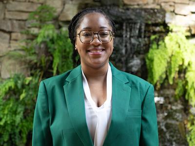 Promise McCoy, 2026-2027 State 4-H Council Secretary-Treasurer, wearing a green jacket and white blouse, standing in front of a stone wall with greenery and a waterfall.