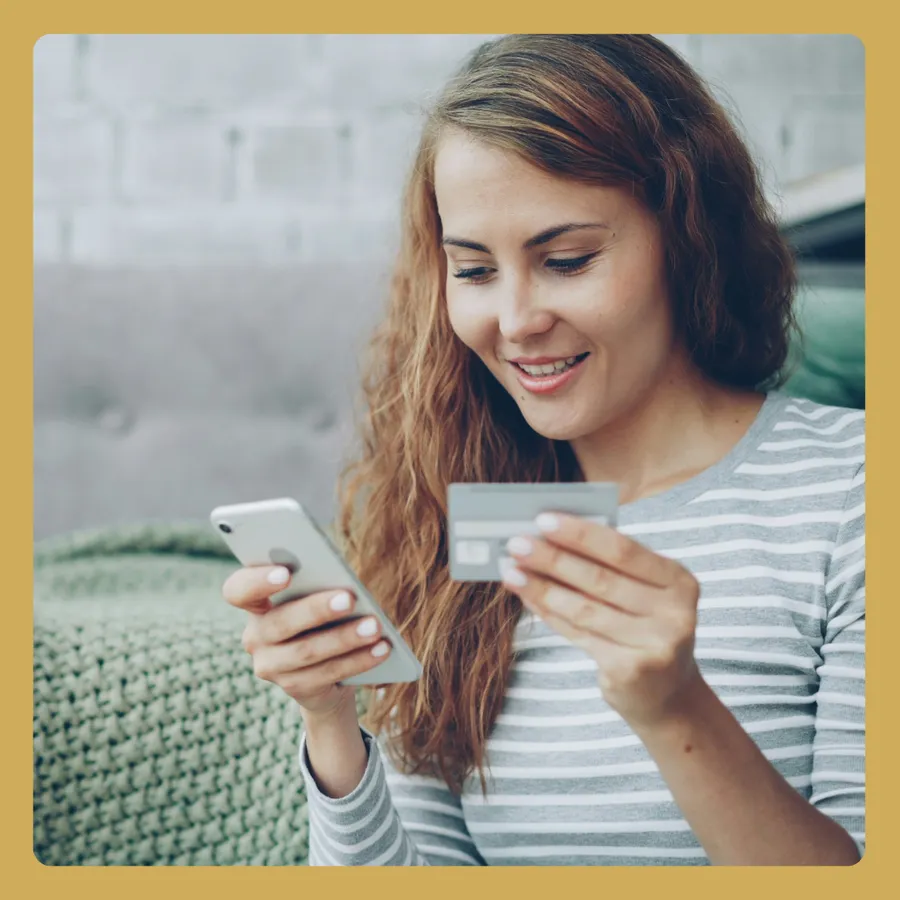 A woman looking at her phone in one hand and holding a credit or debit card in the other