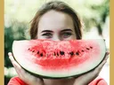 A woman holding a watermelon slice in front of their face with eyes visible above it.