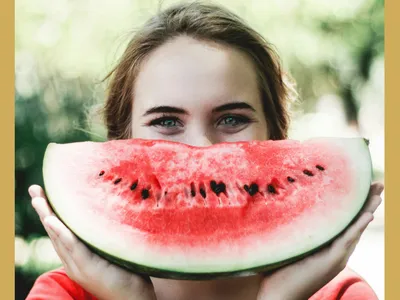 A woman holding a watermelon slice in front of their face with eyes visible above it.