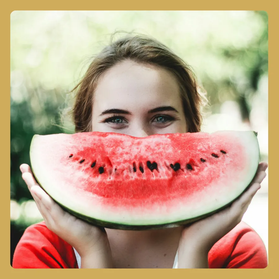 A woman holding a watermelon slice in front of their face with eyes visible above it.