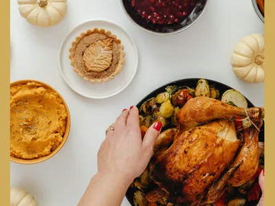 hands placing a holiday turkey on a table with cranberry sauce, mashed sweet potatoes a pie and small decorative pumpkins