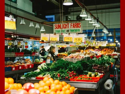A colorful indoor market displaying fresh fruit and vegetables, with a sign reading "SUNLIGHT FARMS." A woman in a striped sweater is browsing the produce.